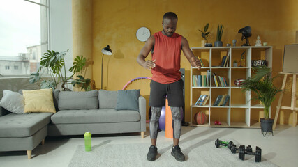 Black sporting muscular young man warming up ahead workout, bouncing jumps exercising aerobics inside apartment. Sport, activity, healthy life. Fitness.