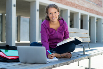 Lachende Studentin lernt vor der Universität mit Buch und Computer