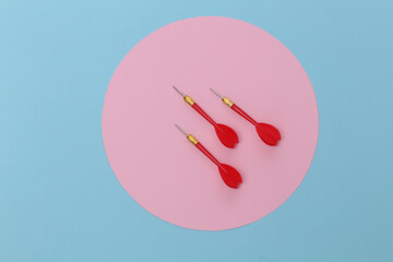 Three red plastic darts with metal tip on blue pastel background with pink circle. Top view. Flat lay. Minimalism