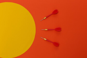 Three red plastic darts with metal tip on orange background with orange circle. Top view. Flat lay. Copy space