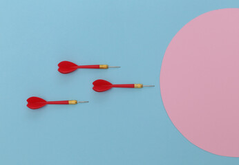 Three red plastic darts with metal tip on blue pastel background with pink circle. Top view. Flat lay. Copy space