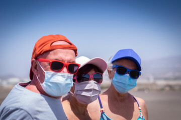Portrait of three friends senior people enjoying together  the beach under sun wearing face mask due to coronavirus - active retired elderly new normal concept