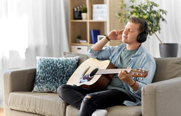 leisure, music and people concept - young man or musician in headphones playing guitar sitting on sofa at home
