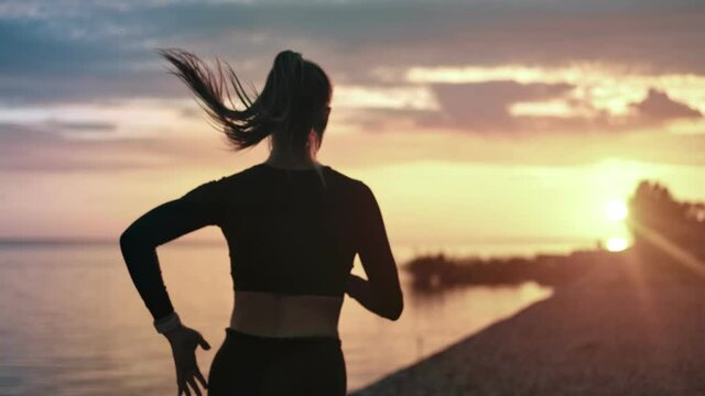Back view sportswoman running at seashore with dramatic sky and sunshine. Medium shot on RED camera
