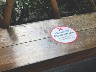 The white and red sticker labeled "Please respect social distancing" is placed on a long bench in the park. Notice for be careful During the COVID 19 epidemic.