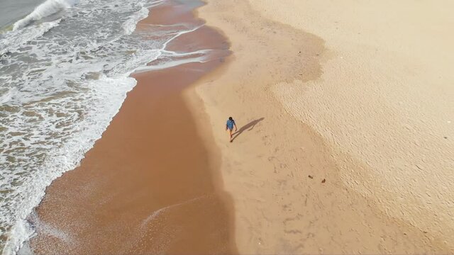 Top-down Aerial Follow shot of a 27-year-old young Indian male walking alone in the seashore by himself shot with a drone in 4k. An empty beach in the coastal town of Pondicherry, India