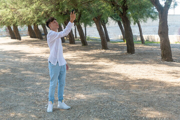 During the day a young italian man with short hair jeans pants and white cotton shirt and white gym shoes between trees in the park at the seaside uses or plays with his phone standing in the blurred 