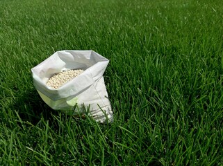 Fertilizer for grass, lawn, meadow in a bag of white granules on a background of green grass. Close up of mineral fertilizer granules used on grass lawns and gardens to maintain health and growth. - obrazy, fototapety, plakaty