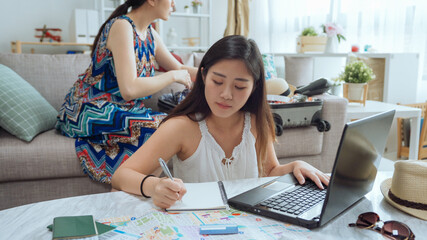 Young woman planning vacation trip and searching information on internet making note on notebook. attractive asian chinese female friend sitting on sofa packing suitcase. two girls on summer trip