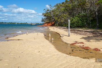 View past sandy shore with tidal creek, red rock and trees to blue sea with boat and blue sky with...