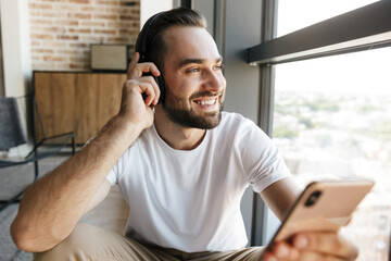 Image of young cheerful man using headphones and mobile phone