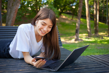 Remote working, education and technology concept. Young smiling beautiful student girl in the city park, wearing white t-shirt, in wireless earphones, using smartphone with laptop preparing for exams.