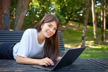 Remote working, education concept. Young smiling pretty student woman, lying on the bench in park, wearing wireless earphones, taking notes and making schedule on laptop preparing for exams. 