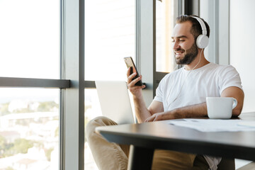 Image of smiling man using laptop and cellphone while sitting at table