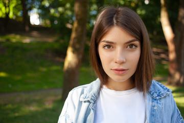Fototapeta premium Close-up portrait European young brunette student female outdoor, wearing white t-shirt and denim jacket, posing against nature background in the summer park and looking to the camera. Free Space.