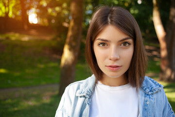 Obraz premium Close-up portrait European young brunette student girl outdoor wearing white t-shirt and denim jacket, posing against nature background in the park and looking to the camera.