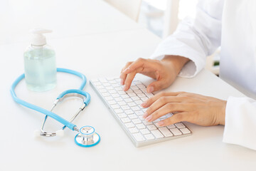 Closeup of female hands typing on computer keyboard, gel hand sanitizer and stethoscope on office desk. Online medical consultation and virus protection concept.
