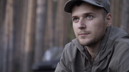 Portrait of young adult male tourist sits on log near fire in forest. Human looks at forest fire on nature. Cooking dinner on wild hike. Outdoor travel. Close up shot