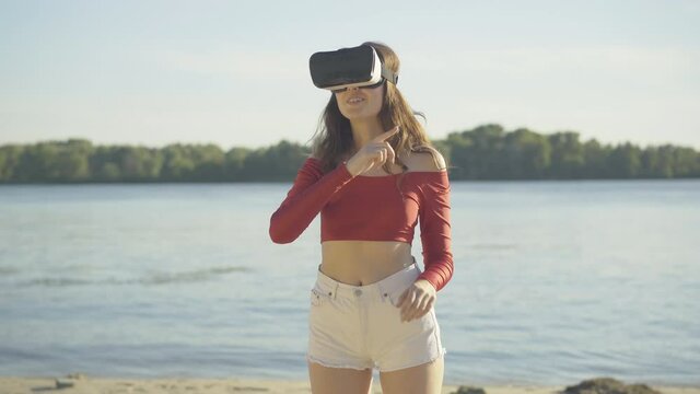 Middle shot of positive young woman in VR-googles moving hands and smiling. Beautiful brunette Caucasian lady resting in virtual reality standing on river beach on sunny day. Modern technologies.