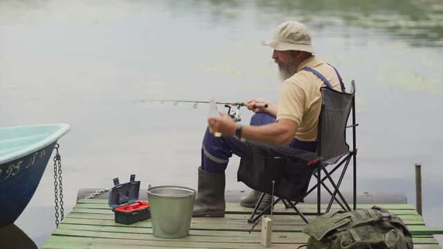Lockdown medium side view shot of bearded old man in bucket hat fishing and drinking lemonade or beer sitting on chair on wooden pier over lake