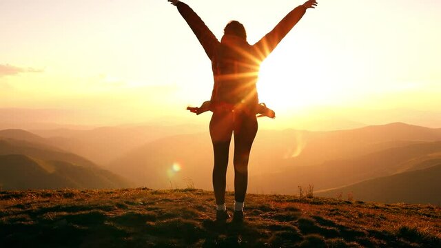 Camera moves around young woman standing victoriously on top of the mountain at sunset. Hiker girl raises arms into air, happy and drunk on life, youth and happiness
