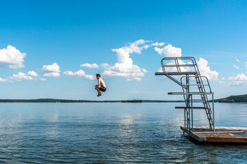 Side view of a teenage male jump diving from a diving tower with blue sky and horizon in the background.