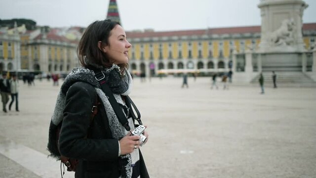 Inspired tourist girl wearing overcoat and scarf, standing on European square, using photo camera, taking pictures, admiring landmarks. Dolly shot. Travel concept