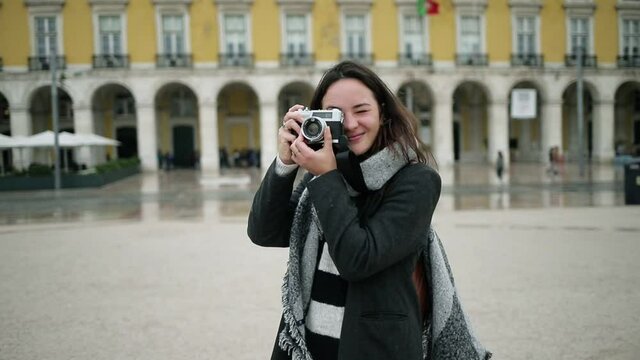 Cheerful tourist girl wearing overcoat and scarf, standing on European square, using photo camera, taking pictures, admiring landmarks. Dolly shot. Travel concept