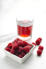 Vertical image.Glass of raspberry drink, square bowl of raspberries o the white marble table