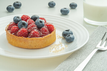 Blueberry and raspberry tartlets served on a transparent plate with brown sugar. Glass of milk in the sun on a white background.
