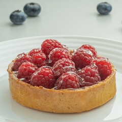 Raspberry tartlets served on a transparent plate with brown sugar. Glass of milk in the sun on a white background.