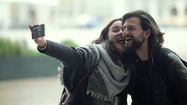 Happy cute young couple wearing overcoats, standing in city, taking self picture on rear smartphone camera. Girl posing and kissing boyfriend. Medium shot. Leisure concept