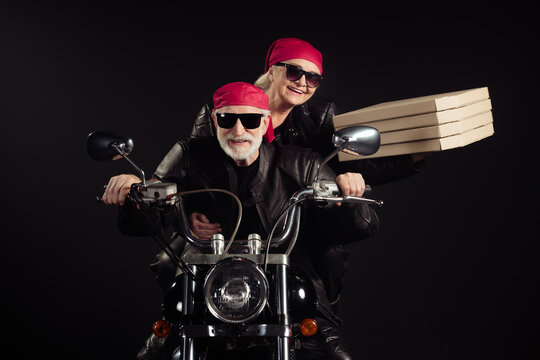 Photo of two cool old bikers white hair man lady couple drive vintage chopper travel festival bring takeaway pizza wear rocker leather jacket outfit bandana isolated black color background
