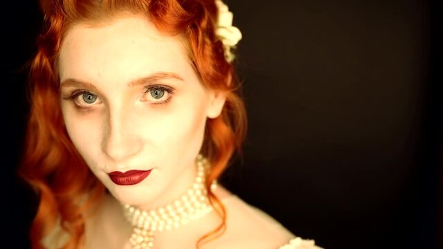 Young vampire woman with long curly hair, pale skin in a white dress on a black background. A beautiful redhead model with red lips. Outfit for halloween