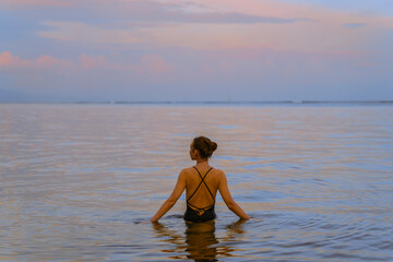 A beautiful girl bathes in the warm sea at sunset. The calm sea reflects the sunset colors of the sky.