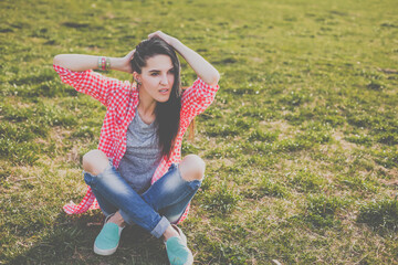 cute hipster woman sitting on the meadow. Looking at camera. Positive human emotion facial expression body language, concept of funny girl. Dressed in a gray t-shirt, and a shirt in a red cage