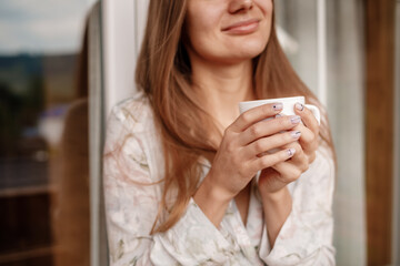 Young female standing after taking a shower in the morning on balcony of the hotel. holding a cup of coffee or tea in her hands. Looking outside nature forest and Mountain