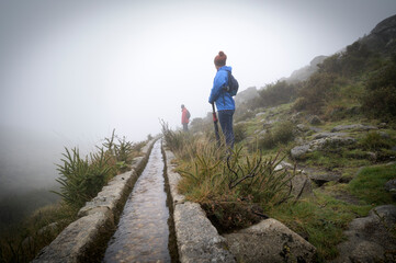 Two people hiking in the mountains in a foggy and rainy day. Girl with blue jacket and umbrella and man with red jacket. - obrazy, fototapety, plakaty