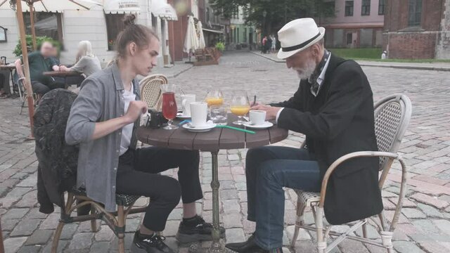 mature father writes and shows paper to adult son at table with fresh drinks in street cafe on old city square