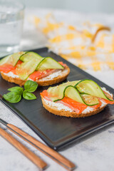 Open sandwiches with trout and cream cheese with cucumber and herbs on a dark plate - a tray on a gray table on a bright sunny day