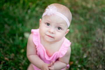 portrait of a baby girl 10 months old sitting on the grass in summer in a pink bodysuit and looking up, walking in the fresh air