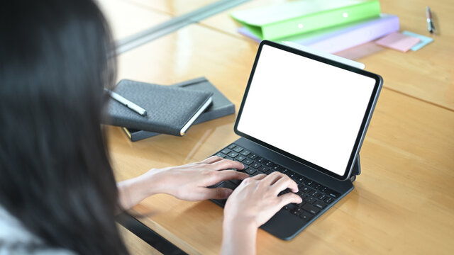 A cropped image of a woman is using a white blank screen computer tablet at the wooden table.
