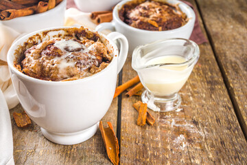 Cinnabon cake in mug. Fast simple microwave dessert idea, background for recipe. Cinnamon roll mugcake, with sugar and cream cheese topping, in different mugs, with cinnamon sticks on wooden backgroun