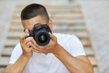A young man is sitting outdoors with a camera and wants to take a photo.