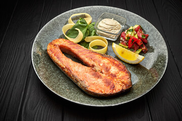 Salmon steak with vegetables, lemon and sauce, on a black wooden background