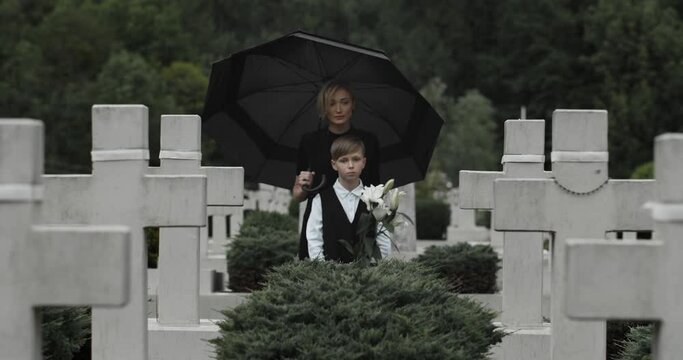 Woman in dark clothes hugging her son with white lily flower at cemetery. Boy looking to camera while standing with widow in row of stone crosses under umbrella. Concept of memorial day