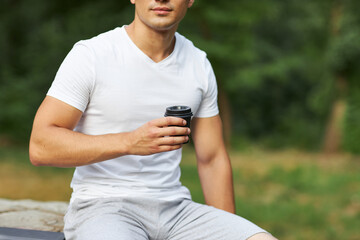 A young man sits in the park on a large stump with coffee in his hands.