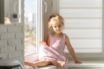 Fototapeta na wymiar beautiful little girl in a pink dress sits on the window on a sunny day