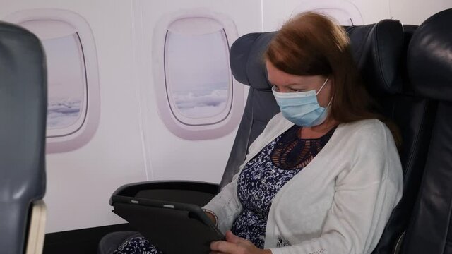 4K: Female Caucasian Airplane Passenger using a Digital Tablet wearing Face mask on plane journey. Air Travel. Stock Video Clip Footage