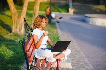 Beautiful smiling young african woman sitting on a bench outdoors, working on laptop computer